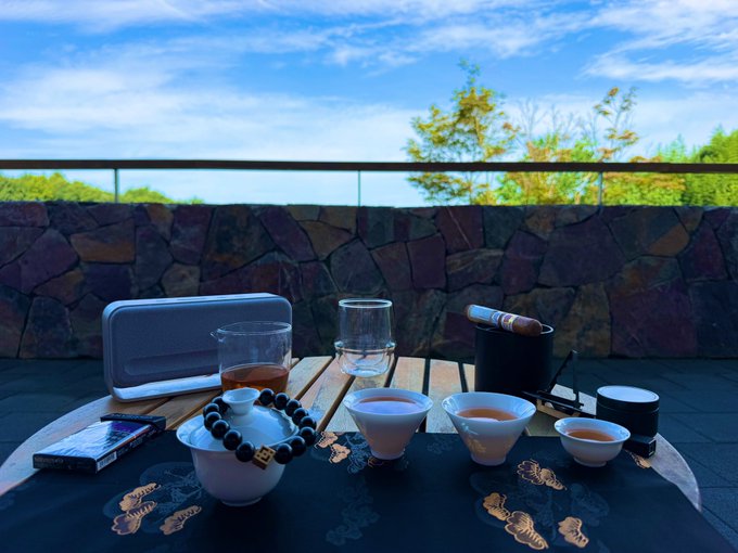 A traditional Chinese tea ceremony setup is arranged on an outdoor table, featuring a white gaiwan teapot with beaded bracelet, small teacups filled with brewed tea, a glass pitcher, and what appears to be a cigar in a holder, all set against a scenic backdrop of sky and trees. The relaxed, contemplative atmosphere of enjoying tea in a serene setting reflects the post's casual, reflective tone as the author notes Bitcoin's quiet climb back to $64,000 while pondering whether others are profiting from US stocks.
