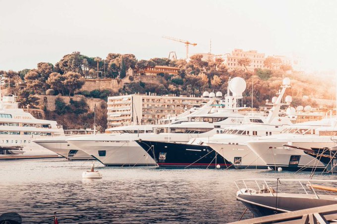 A scenic view of a marina with multiple luxury yachts docked, set against a backdrop of hillside buildings and trees under a warm sky, with a small buoy floating in the foreground.