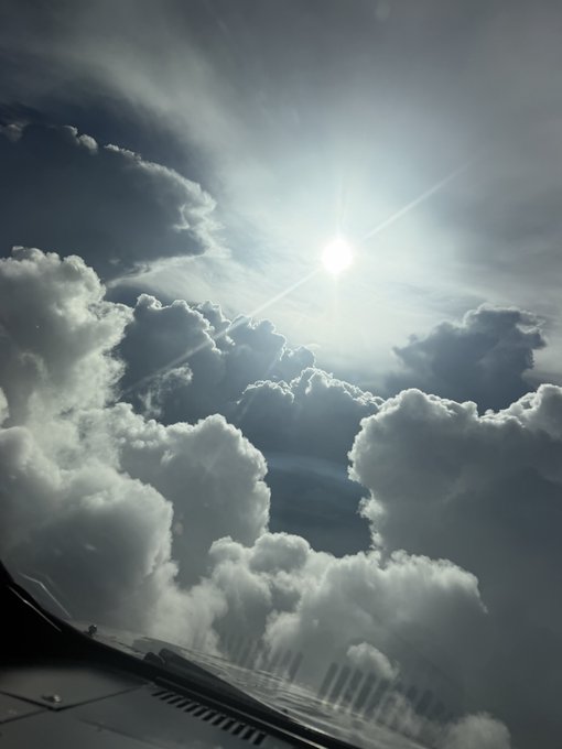 A view from an airplane cockpit window showing bright sunlight shining through dense, billowing white clouds.
