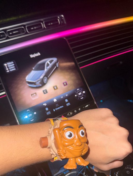 A wrist adorned with a quirky orange-faced character watch is shown in front of a modern car's infotainment display, where the name "MajaLola" appears on screen above a car graphic. The post humorously suggests hiding a cryptocurrency private key behind the watch face, implying the tokens' future value could exceed that of a luxury Richard Mille timepiece.