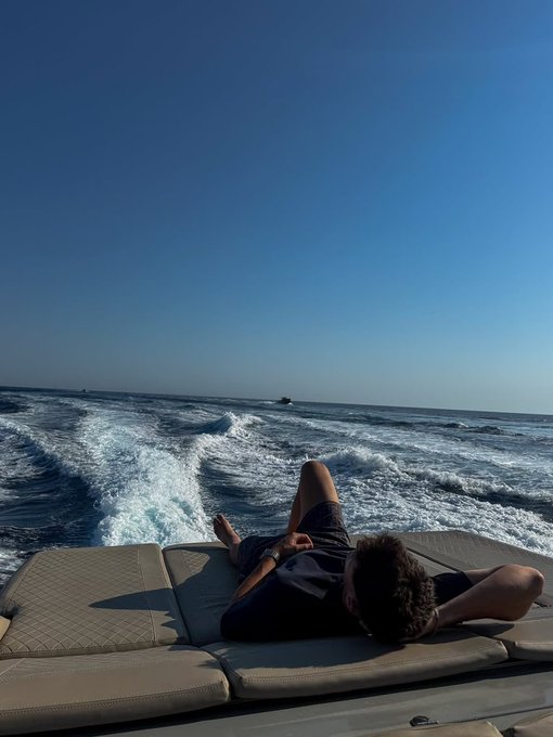 A person relaxes on a cushioned boat deck, lying back while looking out at the ocean wake trailing behind them under a clear blue sky.
