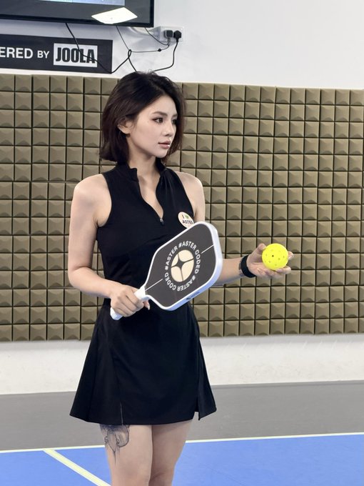 A woman in a black athletic dress stands on a blue pickleball court, holding a paddle branded "Aster Master Coded" along with a yellow ball, with a "Powered by JOOLA" sign visible on the wall behind her. The paddle's branding directly ties to the post's #AsterCoded hashtag, reflecting a passion for the sport of pickleball.