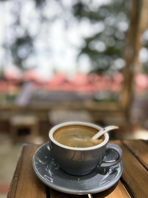 A close-up of a small cup of coffee with a spoon on a saucer sitting on a wooden table, with a blurred outdoor background.