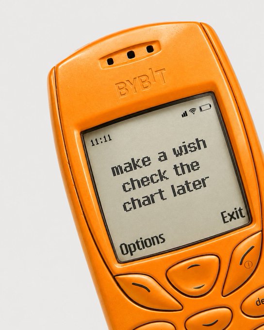 An orange "BYBIT" branded phone displays the message "make a wish check the chart later" alongside "Options" and "Exit" prompts, fitting the "Manifest it in the comments" theme by playfully suggesting crypto traders set intentions and revisit their trading charts afterward.
