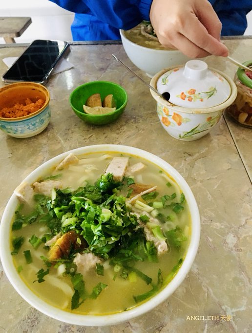 A bowl of soup topped with herbs sits on a table alongside small condiment dishes, with a person reaching toward a lidded cup.