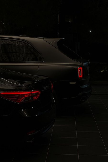 A low-light photograph showing the rear ends of two dark luxury sedans parked on a tiled surface at night, with their distinctive red taillights illuminated.