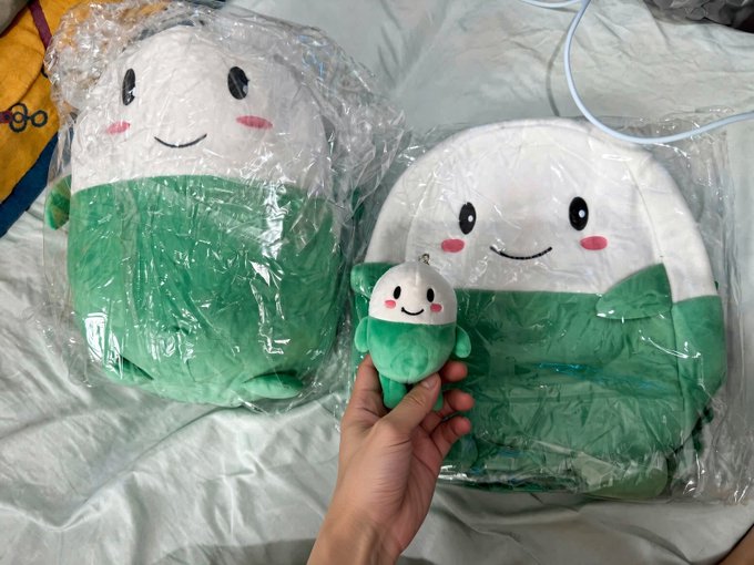 A hand holding a small green and white plush character in front of two larger plush pillows featuring similar smiling designs, still in their plastic packaging on a bed.
