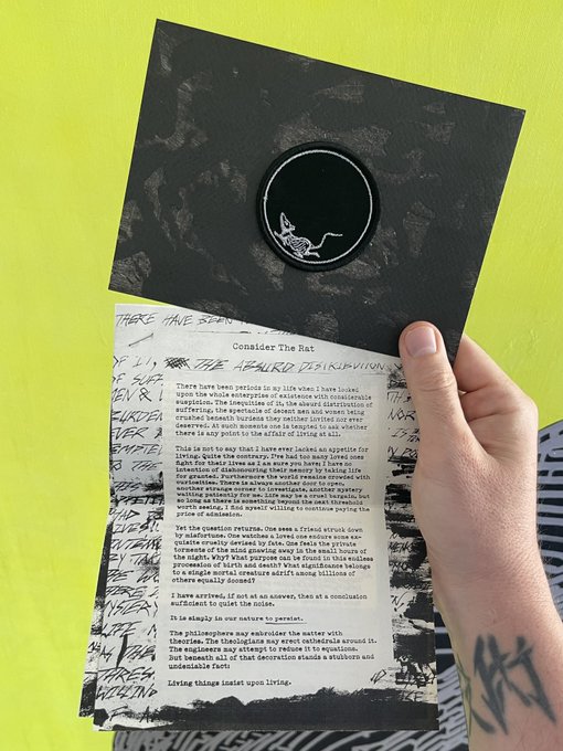 A hand holds a black zine titled "Consider The Rat" opened to a page of typed text, set against a yellow background with handwritten scribbles visible underneath.
