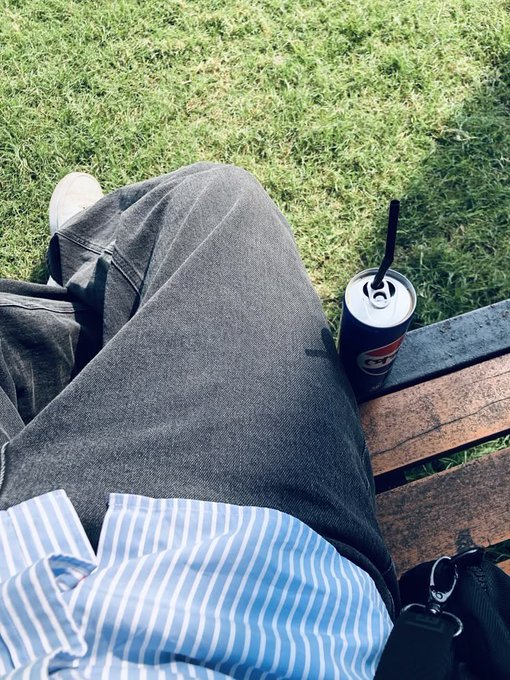 A first-person perspective shows someone relaxing on a wooden bench with their legs crossed, wearing grey jeans and a blue and white striped shirt, with a soda can resting beside them on grass.