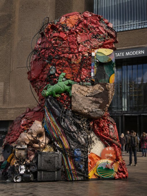 A large mixed-media sculpture of a human head, composed of red debris, wires, and found objects, sits outside the Tate Modern art museum in London.