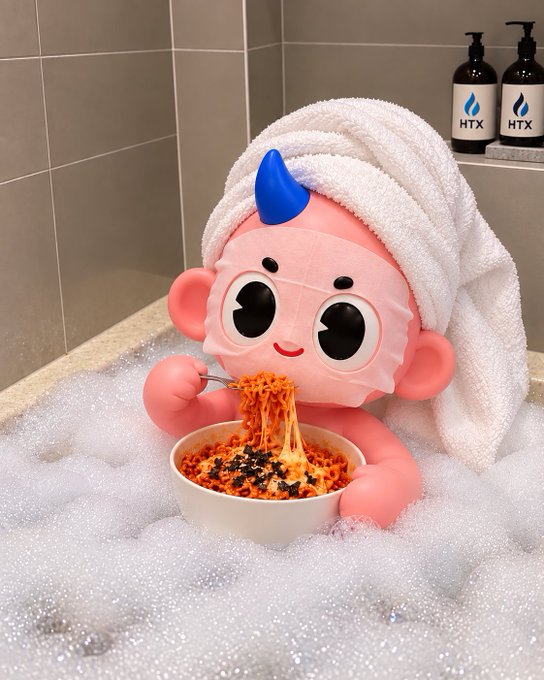 A pink character figurine with a blue horn, wearing a towel on its head, sits in a bubble bath eating a bowl of noodles.