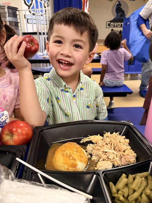 A smiling young boy holds up a red apple in a school cafeteria, with a lunch tray containing chicken, gravy with a roll, and green beans in front of him.