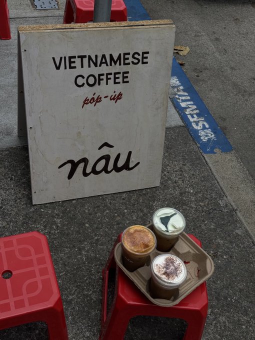 A white A-frame sign advertises 'VIETNAMESE COFFEE' and 'pop-up' with the word 'nâu', sitting next to a red stool holding a tray with three different coffee drinks.