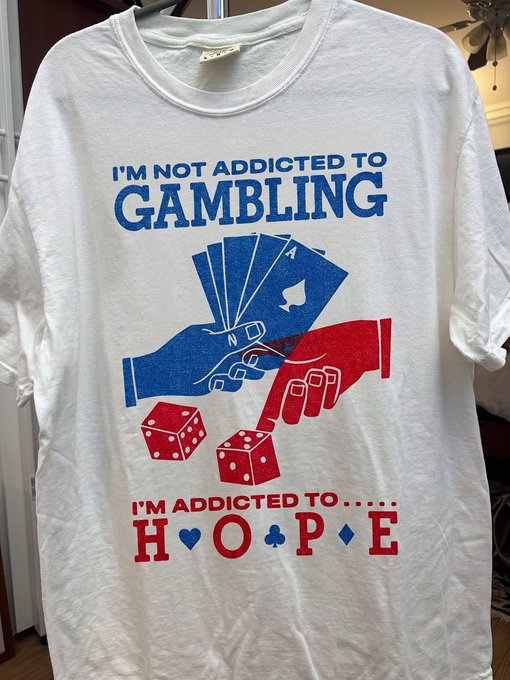 A white t-shirt displayed on a hanger features a graphic design with the text "I'M NOT ADDICTED TO GAMBLING" in blue, "I'M ADDICTED TO.... H♥O♣P♦E" in red (with playing card suit symbols between the letters), and an illustration of two hands holding cards with red dice below.