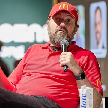 A bearded man wearing glasses and a red McDonald's cap holds a microphone while speaking on stage, with a "holo" branded item visible nearby, representing Cardano founder Charles Hoskinson as he warns that Bitcoin could lose its top cryptocurrency position to quantum computing threats.