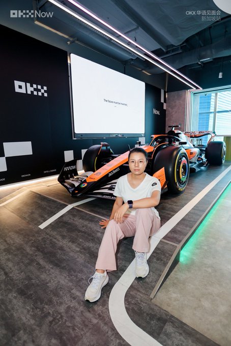 A young woman sits on the floor in front of an OKX-branded McLaren Formula 1 car at the "OKX 华语见面会 新加坡站" (OKX Chinese Meetup Singapore), with the "OKX Now" logo visible on the wall behind her alongside the tagline "The future must be intense."