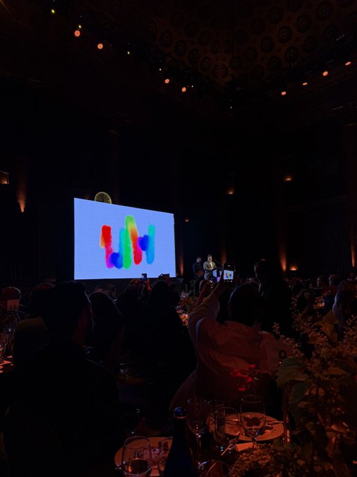A dimly lit conference or awards venue with a large screen displaying a colorful 'W' logo, where a speaker is on stage and audience members are taking photos with their phones.