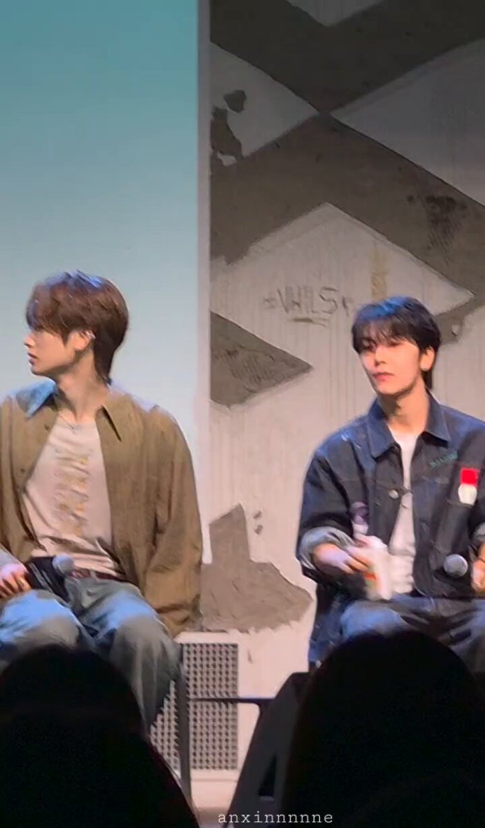 The image shows a stage with two young men sitting next to each other, possibly having a conversation or waiting for their turn to speak at an event. The background features a wall with graffiti art that adds a unique touch to the scene. There is also a microphone and a bottle on the table between them, suggesting they might be discussing something related to the event or sharing refreshments during their interaction.