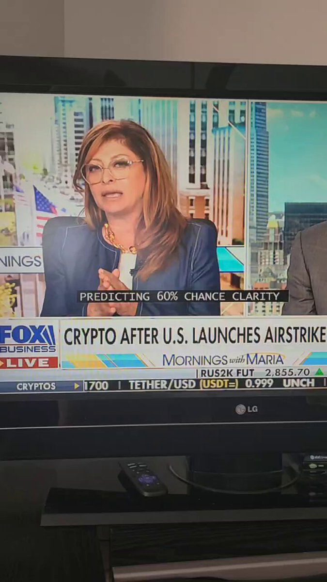 The image shows a television screen displaying a news program with a woman talking about Bitcoin and its impact on the economy. The news anchor is wearing glasses, and she appears to be discussing the potential risks of cryptocurrency. The background features an urban cityscape with tall buildings, suggesting that the story might be related to financial markets or technology trends in the United States.