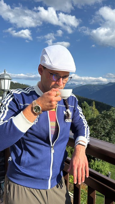 A man wearing a white cap, glasses, and a blue Adidas track jacket drinks from a small white cup while standing on a balcony overlooking a mountainous landscape under a partly cloudy sky.
