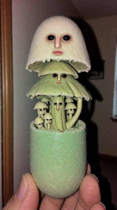 A creepy figurine held in someone's hand depicting a stack of humanoid mushrooms with unsettling human faces, featuring a large white-capped mushroom on top, green layered mushrooms below, and smaller mushrooms emerging from a green base.