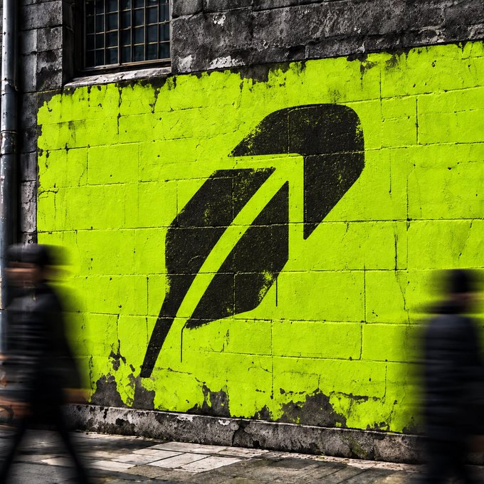 A vibrant neon green wall features a large black "R" logo, while two blurred pedestrians walk past in motion—visually evoking the fast-paced movement of life, fitting the caption "Life feels better on Robinhood."