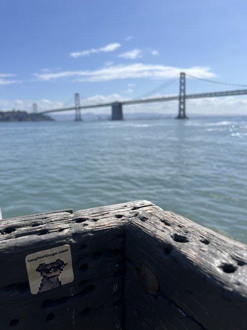 A photograph taken from a wooden pier, looking out over blue water toward a large suspension bridge (resembling the Bay Bridge) under a bright blue sky with scattered clouds; a small pixel-art sticker reading 'Deginathoge.com' is visible on the wood.