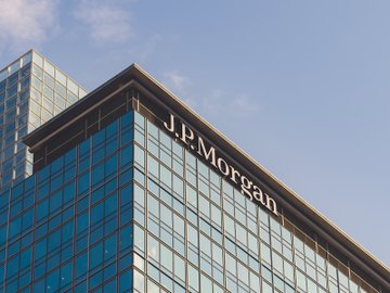 A glass skyscraper prominently displays the "J.P.Morgan" name across its upper facade against a blue sky, visually anchoring the news about JPMorgan analysts commenting on Strategy's $3B cash reserves and their optimistic outlook for Bitcoin.