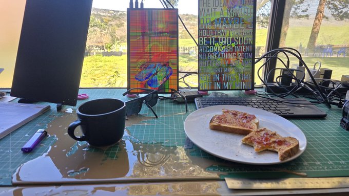 A messy wooden desk holds a spilled black mug of liquid, a plate with two pieces of toast, sunglasses, and a cutting mat, set up as a workspace near a window showing a grassy landscape.