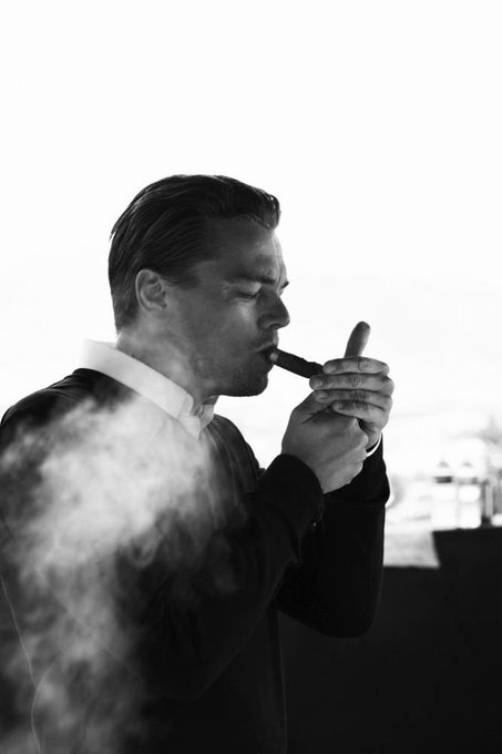 A black and white photograph shows a man in a suit in profile, smoking a cigar with smoke billowing around him.