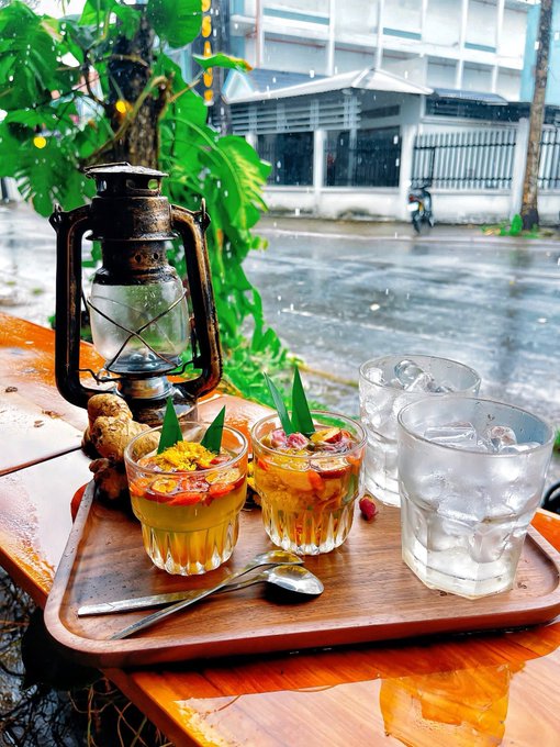 A wooden tray holds two glasses of fruit dessert topped with pandan leaves, a glass of iced water, a vintage lantern, and ginger root, set on a rainy outdoor table with tropical foliage.