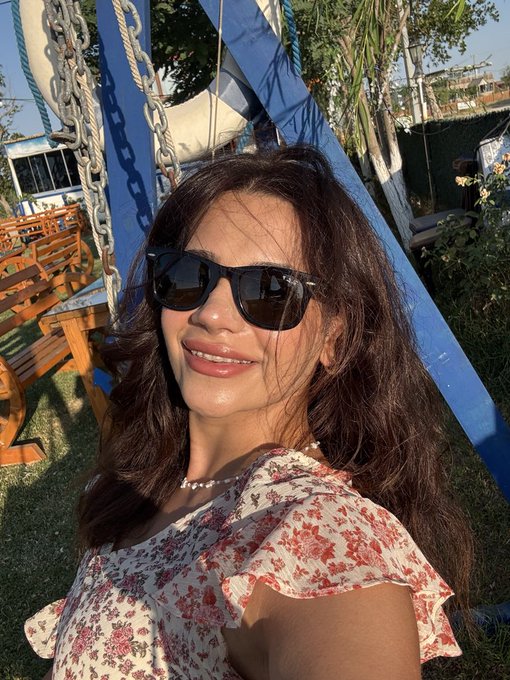 A smiling woman with dark wavy hair and black sunglasses poses for a selfie, wearing a floral-patterned top with a delicate pearl necklace, set against a lush green outdoor backdrop featuring blue structural beams and rope swings that complements her cheerful "GM fam" message.