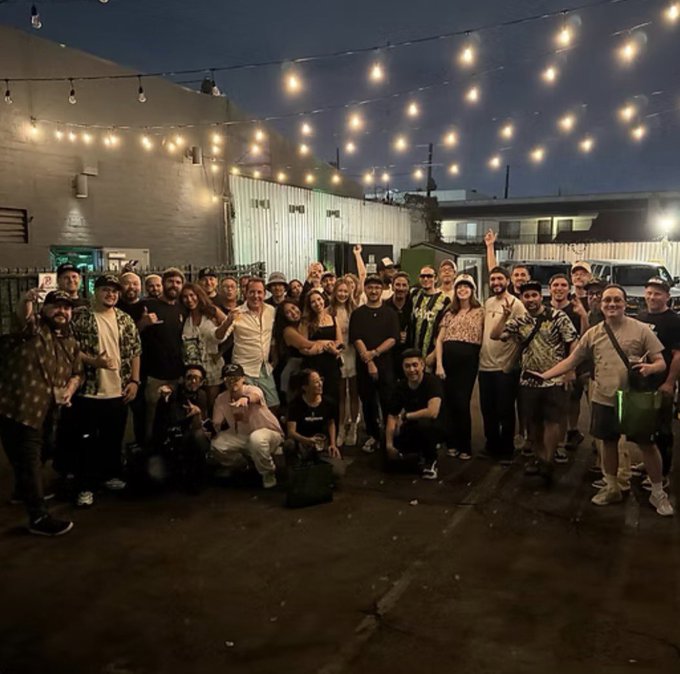 A large group of 150+ Bored Ape Yacht Club (BAYC) members gather in LA at night under string lights to celebrate the club's fifth anniversary, with some attendees wearing Bored Ape-themed shirts and posing together in front of a warehouse.