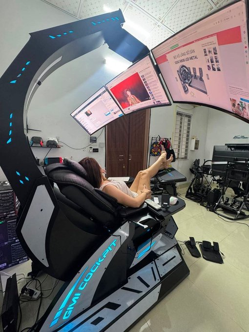 A person reclines in a sleek "GM COCKPIT" gaming cockpit chair in front of a multi-monitor setup, with the screens displaying financial websites, a Bitcoin chart, and YouTube content—matching the post's caption about lounging while analyzing Bitcoin. The futuristic black and blue chair features built-in controls and a pedestal base, suggesting it's a high-end gaming setup worth seeking out for purchase.