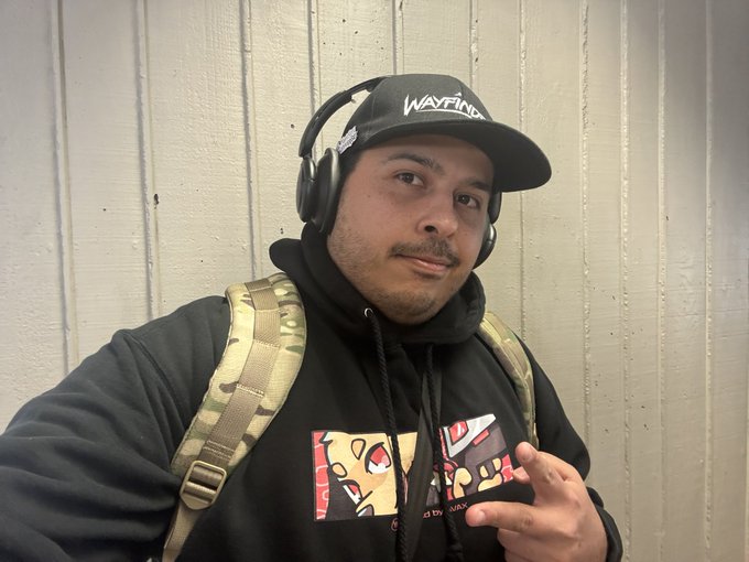 A man wearing a black 'WAYFIND' cap, headphones, and a black hoodie with a Pokemon-themed graphic, carrying a backpack.