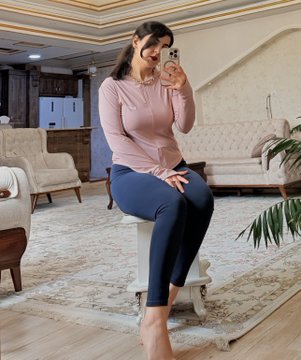 A woman wearing a pink long-sleeve top and blue leggings takes a mirror selfie in a well-furnished living room with elegant white furniture.