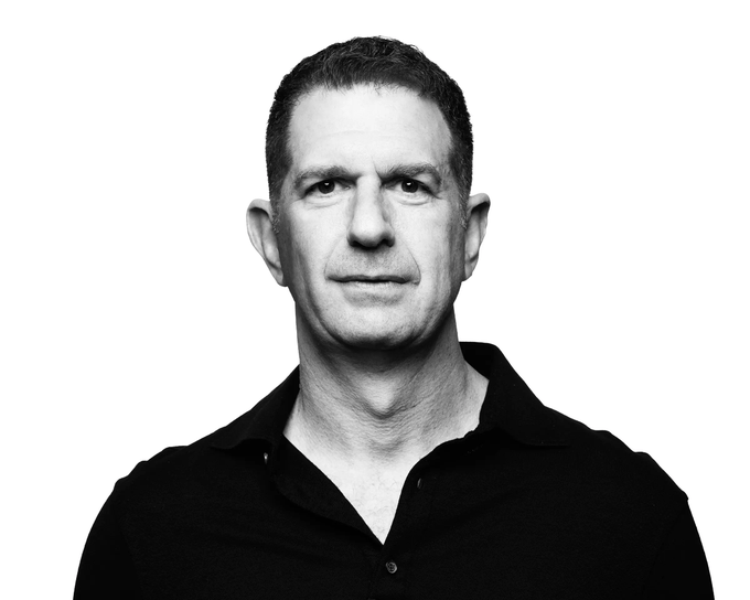 A black-and-white portrait shows a middle-aged man with short hair wearing a black polo shirt, looking directly at the camera against a white background.