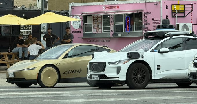 A gold Tesla Cybertruck and a white Jaguar I-Pace self-driving taxi sit parked on a city street, with the former featuring graffiti-style "CYBER" lettering and a solid disc wheel covering. Behind them, a pink food truck labeled "FOOD IS LOVE" with "QUA CHARCUTERIE" signage is visible, while a group of men sit under a yellow umbrella outside a "COFFEE CO" establishment—suggesting the "Brutal mog" caption humorously refers to the Cybertruck's divisive, polarizing design parked next to its futuristic autonomous counterpart.
