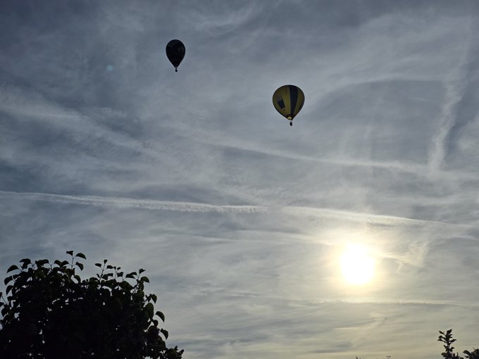 The image features two hot air balloons floating in the sky, with a tree nearby. The sun is setting behind them, creating a beautiful backdrop for this scene. In the background of the photo, there are also some clouds visible, adding depth to the overall composition. The caption "Gm World Happy hump day!!! Anyone buying or have we given up Up Up and awayyyyy!!!!" seems to be related to the hot air balloons in the image, possibly suggesting that someone is celebrating a special occasion or event involving these balloons.