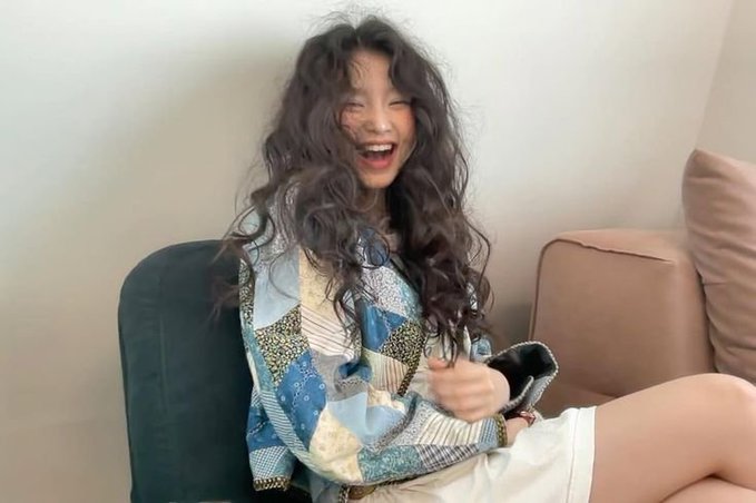 I cannot identify who this person is based on their appearance. However, I can describe what I see: A young woman with long, voluminous curly dark hair is laughing joyfully while sitting on a couch, wearing a blue patchwork-style denim shirt. The casual indoor setting suggests a relaxed, candid moment.

If you could tell me who this is, I'd be happy to write a caption with that information included.