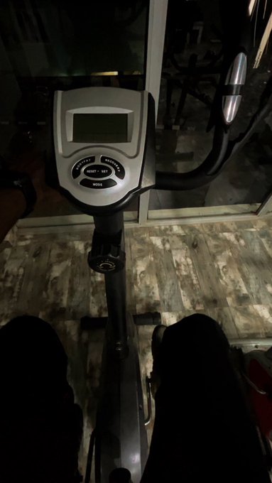 The image shows a person's legs positioned on an exercise bike, with their feet resting on the pedal handles. The person appears to be at the gym or workout area during the night time. A caption below the photo reads "Sunday night at the gym, I’m not your mate. The only grass I touch is weight and some grAss. Ending this week grinding on @wolfswapdotapp amplify contest. Goodnight Yamiis".