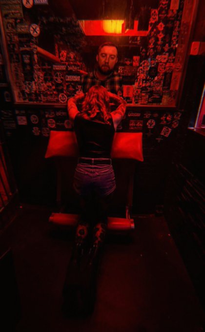 Two people embracing in a dimly lit room with walls covered in stickers and posters, bathed in red light, with one person seated facing away from the camera.