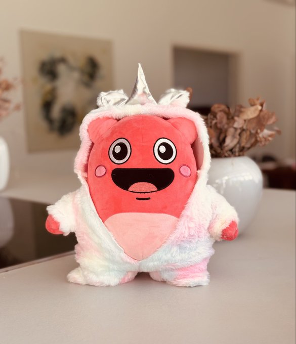 A pink plush toy wearing a rainbow-colored unicorn costume with a silver horn, sitting on a white table in a home interior.