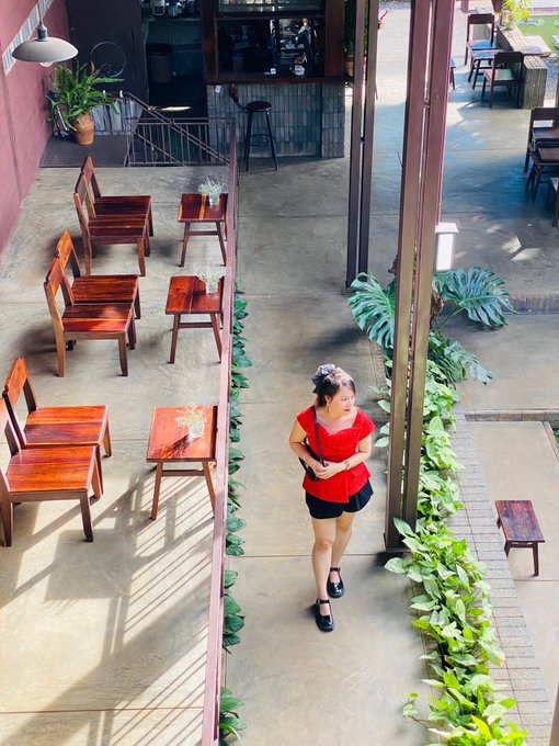 An aerial view captures a woman in a red top and black shorts walking through an outdoor cafe or courtyard area with wooden tables, chairs, and tropical plants.