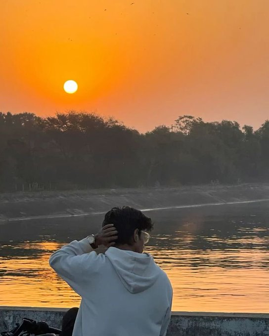 A person in a white hoodie sitting by water at sunset with an orange sky, watching the sun dip below distant trees.