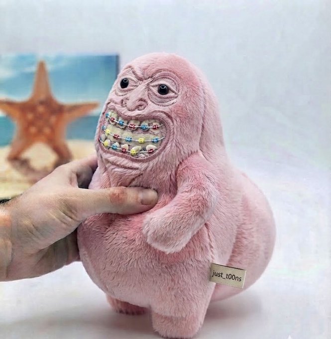 A creepy pink plush toy with a wide grinning mouth full of braces being held by a hand, with a 'just_toons' label.