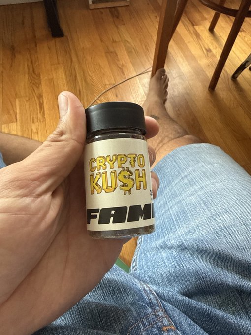 A first-person view of a hand holding a small jar labeled 'CRYPTO KU$H FAME' while sitting on a wooden floor in blue jeans.