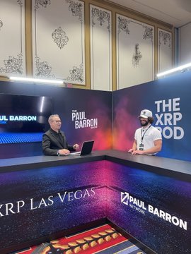 Two men sit across from each other at a podcast studio desk branded with "Paul Barron Network" and "The XRP Pod" signage, with "XRP LAS VEGAS" displayed prominently on the front panel. The setting captures a live interview or recording session during the XRP Las Vegas 2026 event.