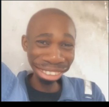A close-up photo of a young smiling Black man wearing a blue jacket against a plain wall background.