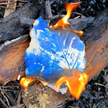 A leaf-shaped object with striking blue and white marbled patterns burns with orange flames atop charred wood.